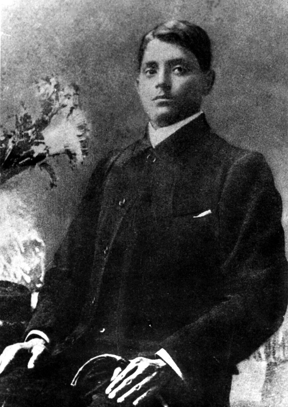 Full seated studio portrait of Jatindranath Mukherjee in a dark buttoned coat, hands resting in his lap, before a painted backdrop.